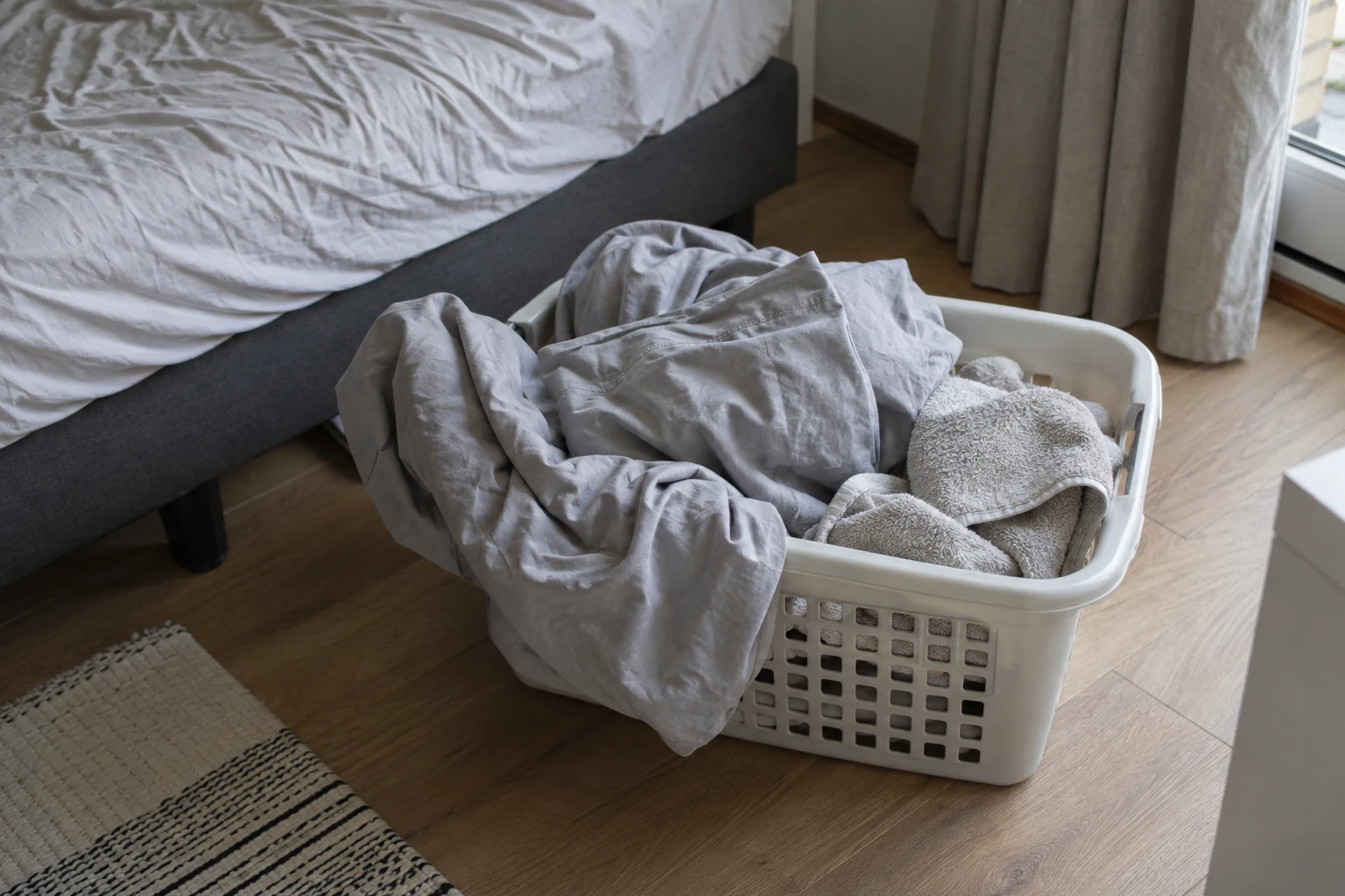Een wasmand met katoenen beddengoed en handdoeken naast een bed