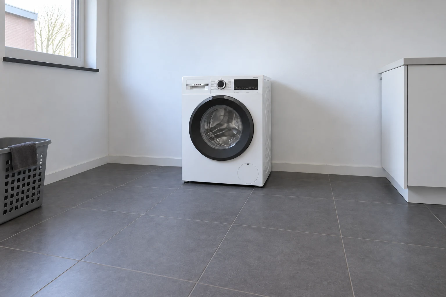 Een witte wasmachine in een lichte wasruimte op een donkere harde vloer, met vrije ruimte ervoor.