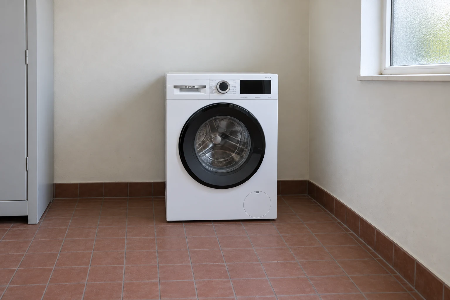 Een gesloten Bosch-wasmachine staat op een tegelvloer in een eenvoudige bijkeuken.